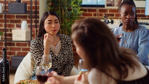 Asian woman at apartment party paying attention to interesting story shared by friend while eating gourmet cheese from charcuterie platter. BIPOC guest listening to group of people chatting photo