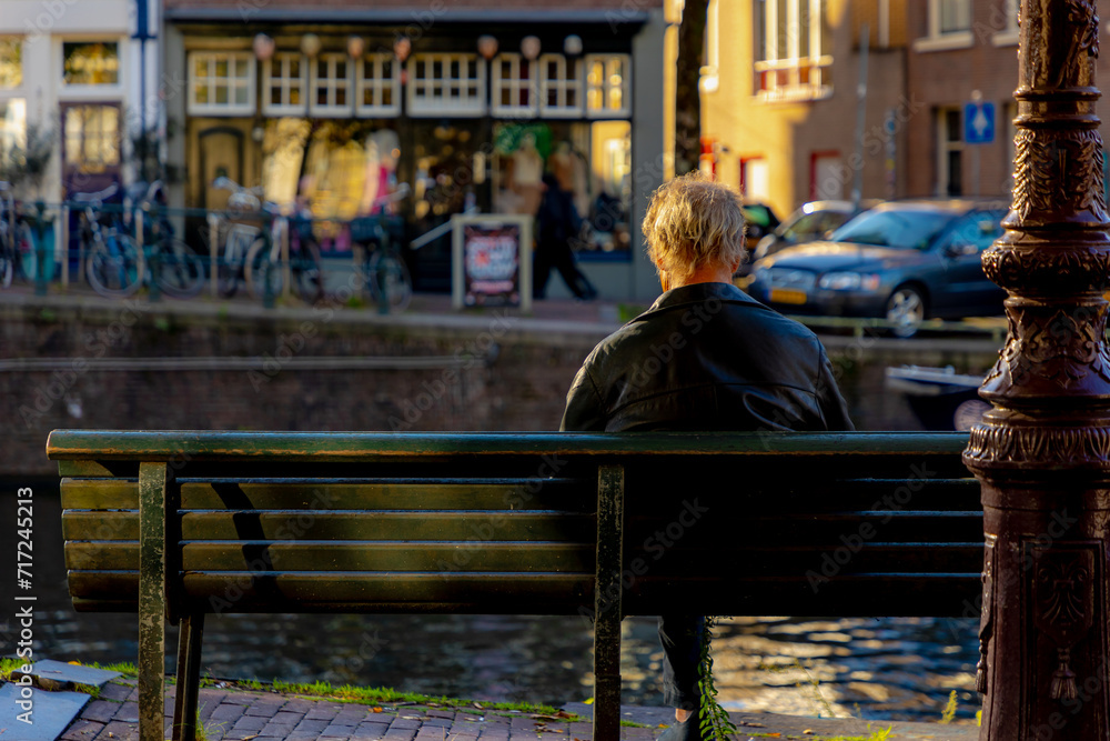 Backside of a old man on wooden bench with blurred view along the canal, The senior guy sitting and relaxing, Hobby and leisure activity of retirement age, Amsterdam capital city of the Netherlands.