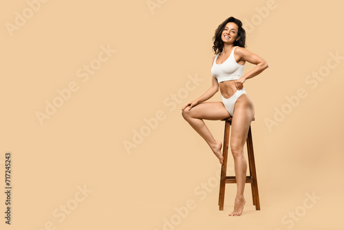 Slender young woman in white underwear sitting on chair over beige background