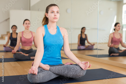Sporty slim girl meditating in yoga position Padmasana during group training at gym.