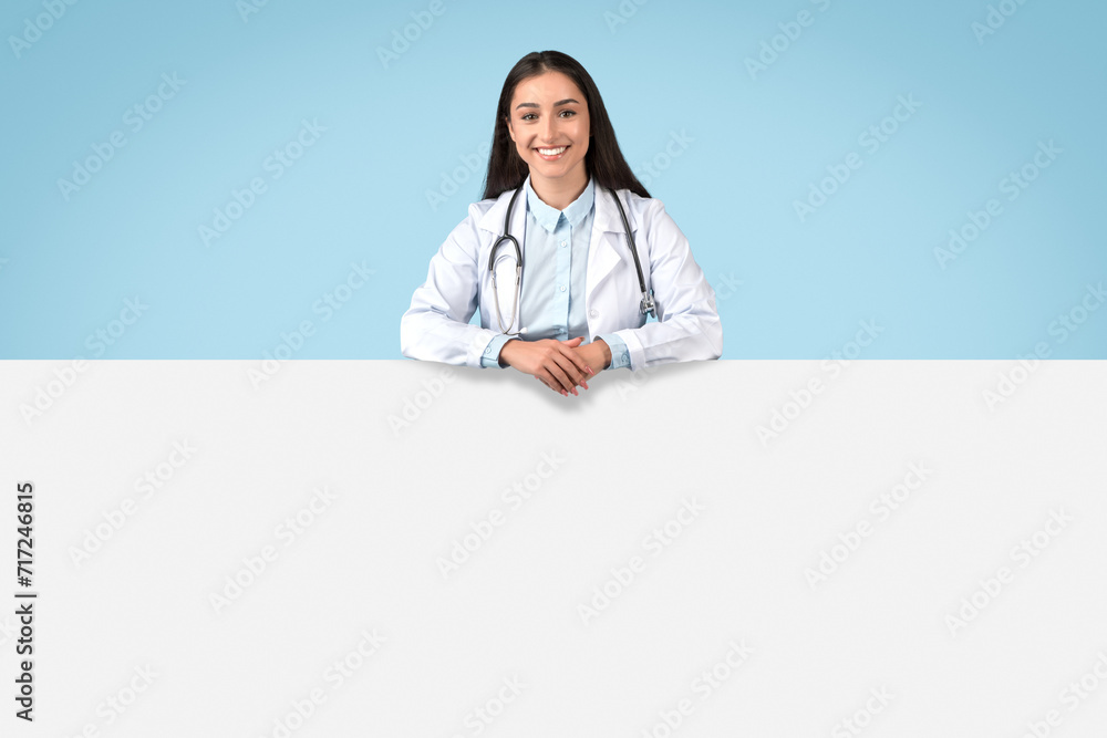 Happy young caucasian lady doctor in white uniform, leaning on white blank placard for medical advertising offers Happy young caucasian lady doctor in white uniform, leaning on white blank placard for medical advertising offers