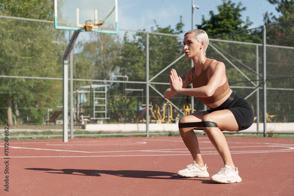 An athletic woman engages in a powerful squat workout with resistance bands at a sunlit basketball court