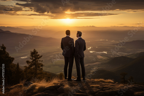 Two Successful Business Partners Sharing a Moment of Triumph on a Hill, Overlooking the Vibrant Colors of the Valley Below as the Sun Sets © aicandy
