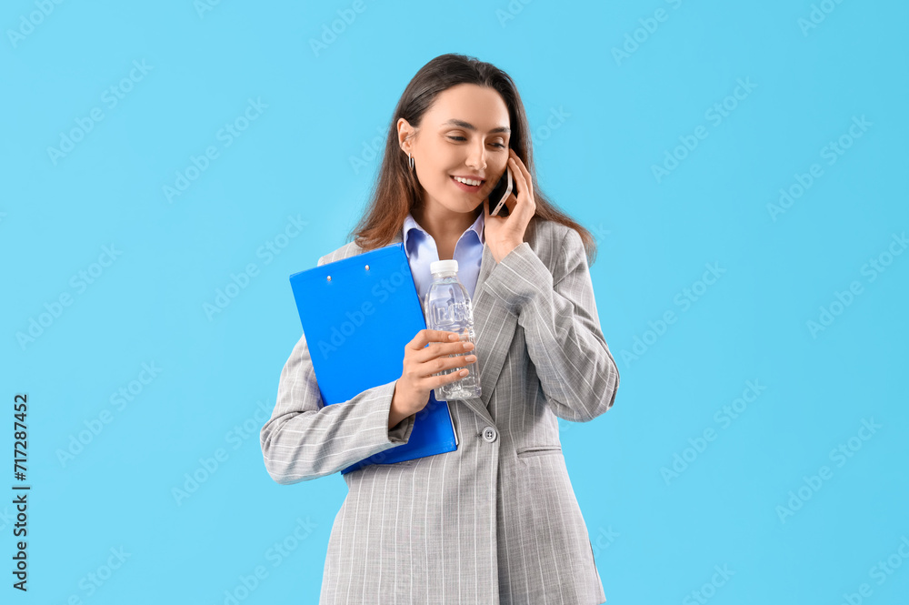 Young businesswoman with clipboard and bottle of water talking by mobile phone on color background