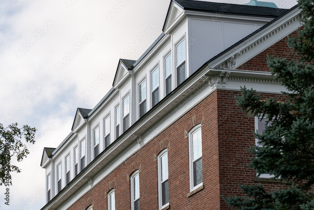 Fototapeta premium The exterior of a large multi-level university brown brick building with rows of multiple glass windows. The shingled white wooden roof has a row of peaked dormers with double-hung glass windows. 