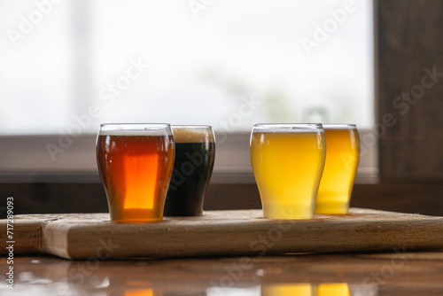 Fototapeta Naklejka Na Ścianę i Meble -  Small curved tulip shaped sample glasses of various ale beers with foam on the top. The samples are stout, red ale, pale ale, and IPA style beer on a wooden flight tray. The samples are on a table.