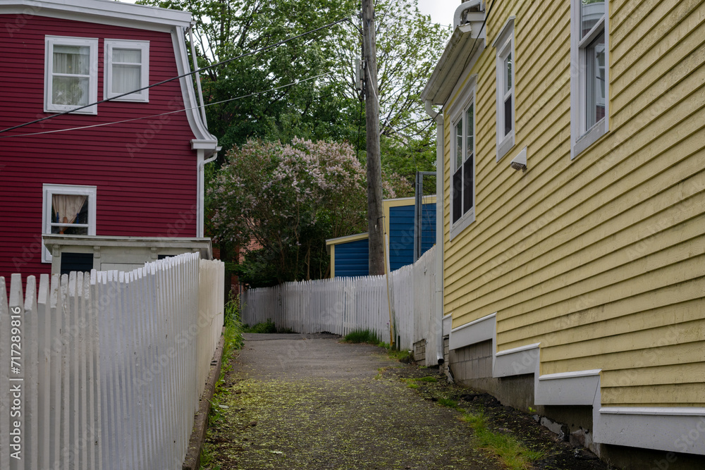 A narrow walkway or pathway behind bright, and colorful houses painted red, blue, and yellow. The wooden historical buildings have narrow clapboard siding and white trim around the windows. 