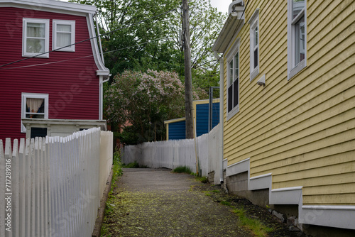 A narrow walkway or pathway behind bright, and colorful houses painted red, blue, and yellow. The wooden historical buildings have narrow clapboard siding and white trim around the windows. 