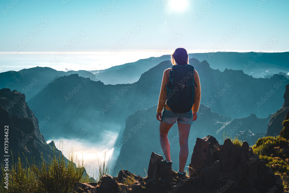 Obraz premium Backpacker tourist stands on the edge of a deep, cloud-covered valley and enjoys the breathtaking panoramic view of the volcanic mountain landscape. Pico do Arieiro, Madeira Island, Portugal, Europe.