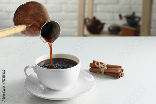 Turkish coffee. Pouring brewed beverage from cezve into cup at white table indoors photo