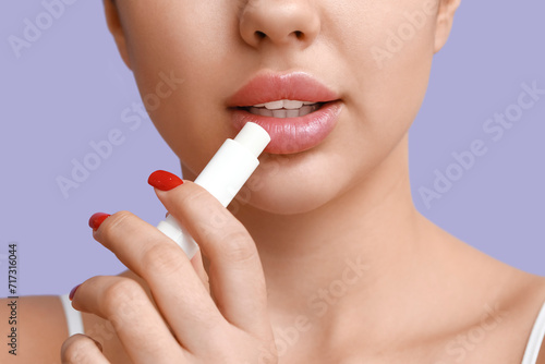 Beautiful young woman applying lip balm on lilac background  closeup