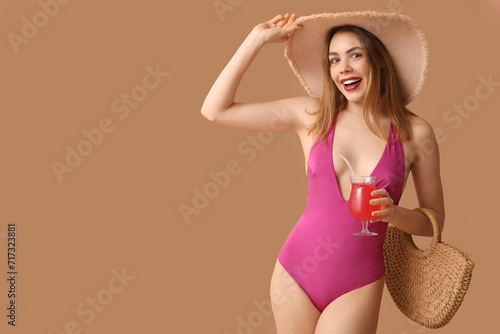 Beautiful young woman in tank swimsuit, with beach bag and cocktail on beige background