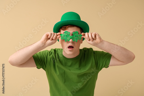Shocked young man in leprechaun's hat with party glasses on beige background. St. Patrick's Day celebration