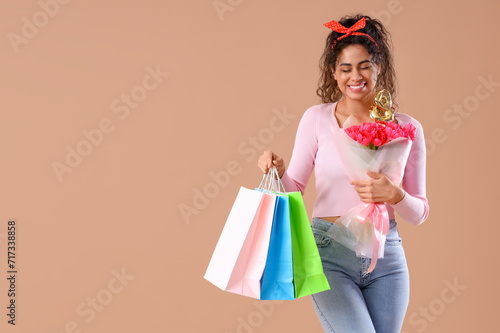 Fashionable African-American woman with bouquet of flowers and shopping bags on beige background. International Women's Day celebration