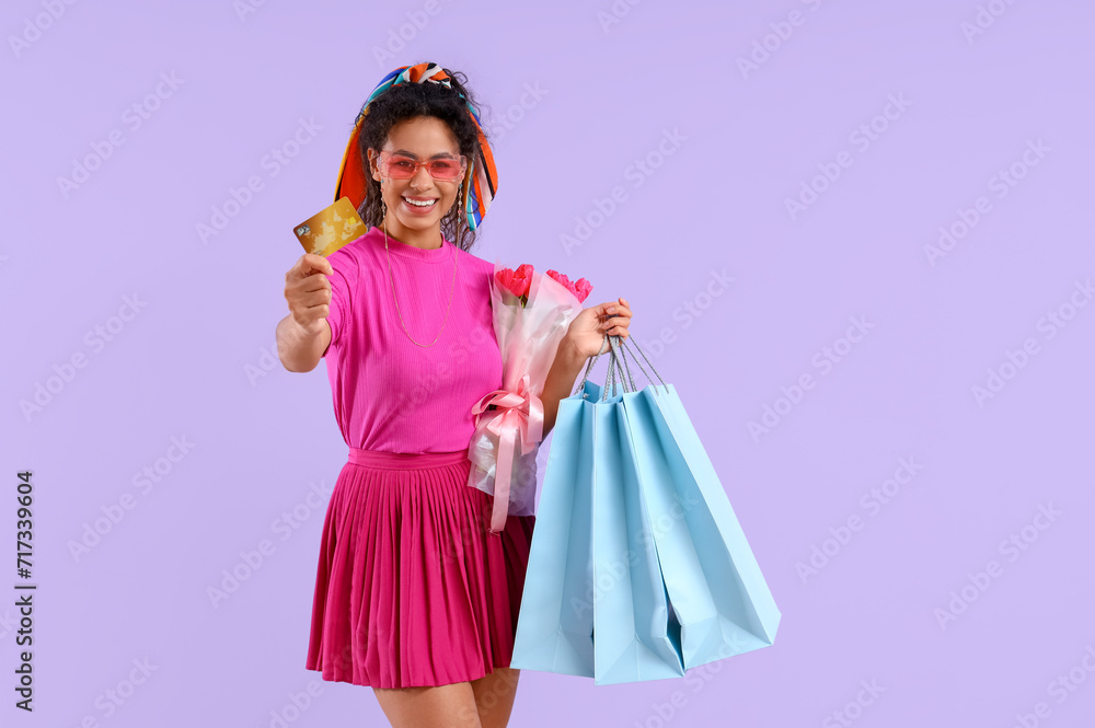 Fashionable African-American woman with credit card, bouquet of flowers and shopping bags on lilac background. International Women's Day celebration