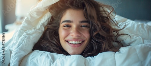 Young woman waking up happily in the morning, smiling while under the blanket in bed.