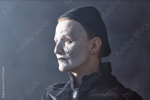 Dramatic headshot portrait of mime artist standing on stage in spotlight and looking away with sad face makeup, copy space photo