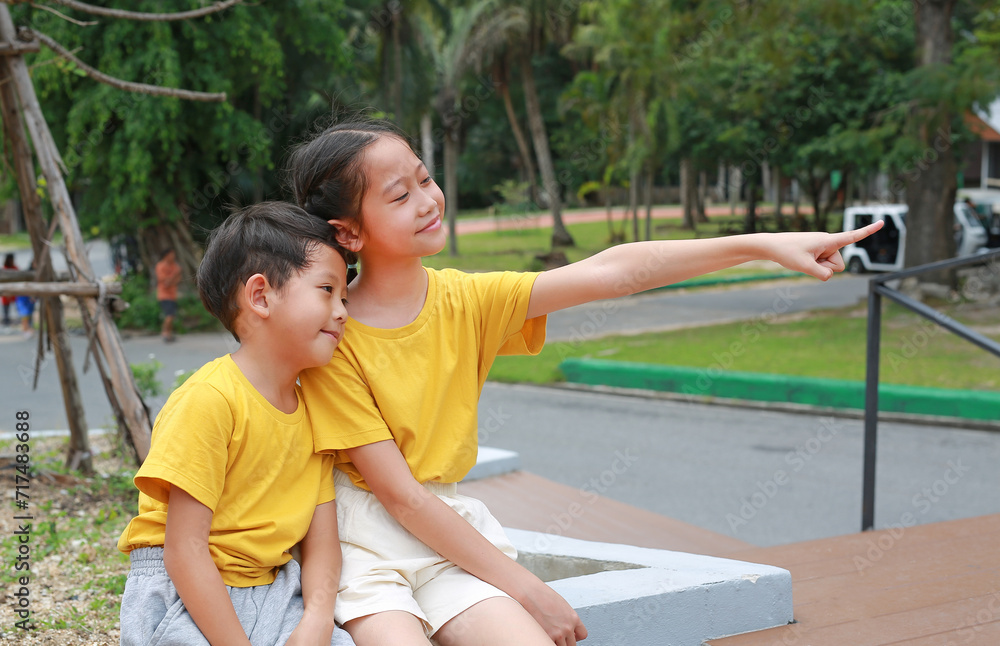 Portrait of Asian boy and girl child are sitting in the park and pointing at something beside them, Children are friends in the garden.