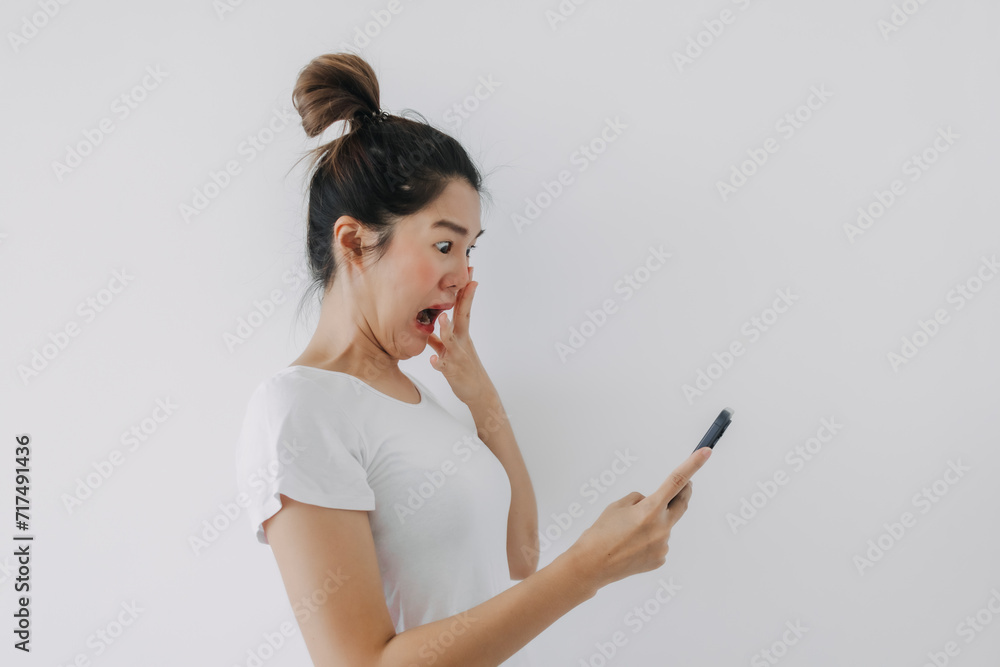 Asian Thai woman holding mobile phone with funny surprised and shocked face, while looking cellphone, standing isolated over white background wall. Asian Thai woman holding mobile phone with funny surprised and shocked face, while looking cellphone, standing isolated over white background wall.