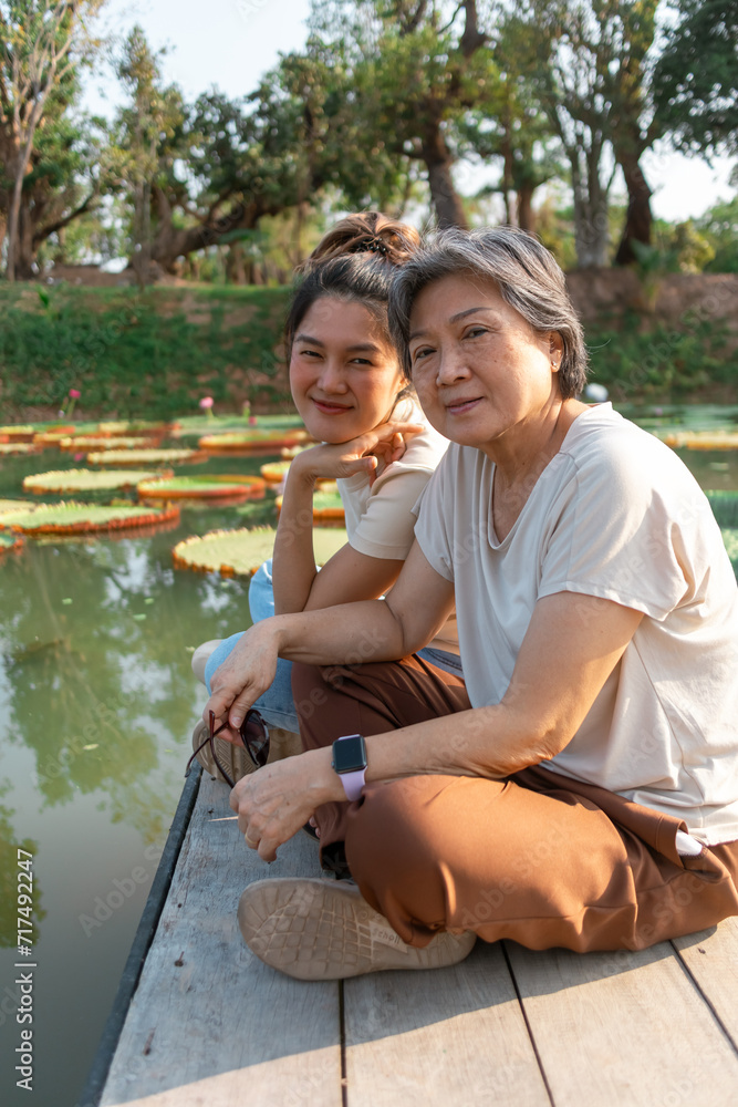 Side view of asian Thai Chinese elder mother and daughter sitting on the bank by lotus flower lake local lagoon, both woman happy smiling to camera, traveling on vacation. Side view of asian Thai Chinese elder mother and daughter sitting on the bank by lotus flower lake local lagoon, both woman happy smiling to camera, traveling on vacation.