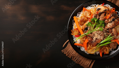 Top view of korean food in black frying pan on dark surface. Cooked asian ingredients in iron skillet on black background with copy space for text. Sautéed noodles, mushrooms and vegetables recipe. photo