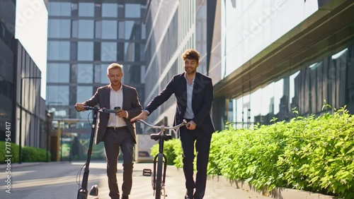 Two businessmen going city downtown. Smiling colleagues talking holding scooter