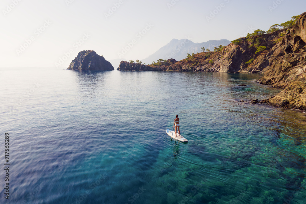 Obraz premium Woman in bikini stand on sup paddle board with paddleboarding passing calm sea with rocky shore. Female enjoy outdoor lifestyle and water active on holiday vacation