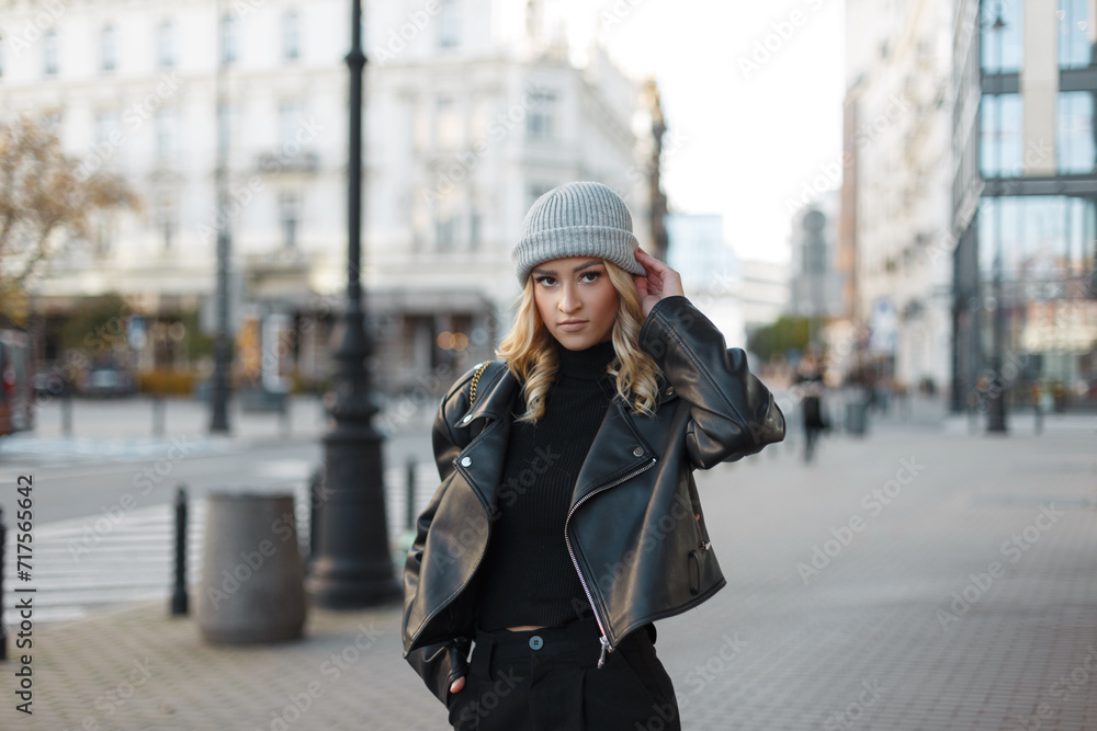 Beautiful young woman model in fashion black rock urban clothing with a hat, black leather jacket and sweater walks in the city