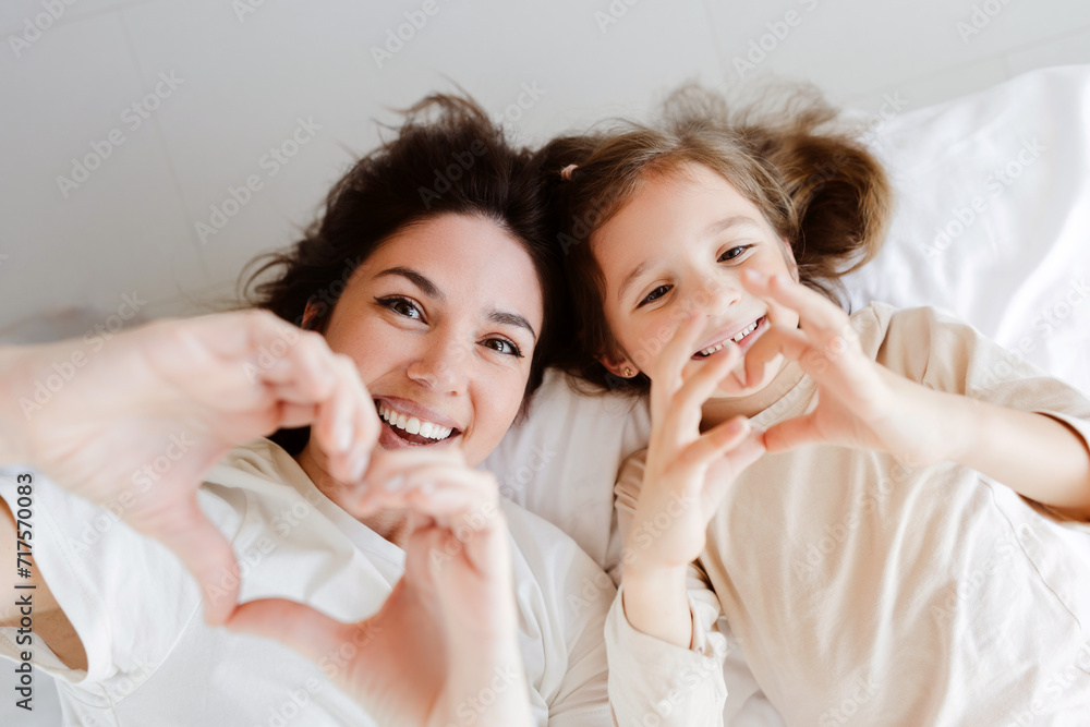 Positive mother and little daughter lying on bed with white bed linen, gesturing showing heart shape Positive mother and little daughter lying on bed with white bed linen, gesturing showing heart shape