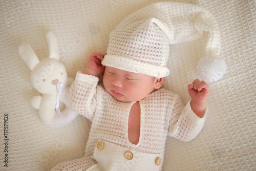 On a white blanket, a cute newborn baby is sleeping with teddy bear. The baby wearing a white suit and hat. Top view