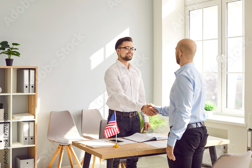 A young attractive man reaching agreement with immigration application and shaking hands with public services worker standing in the office of the US embassy at the desk of official's workplace. photo