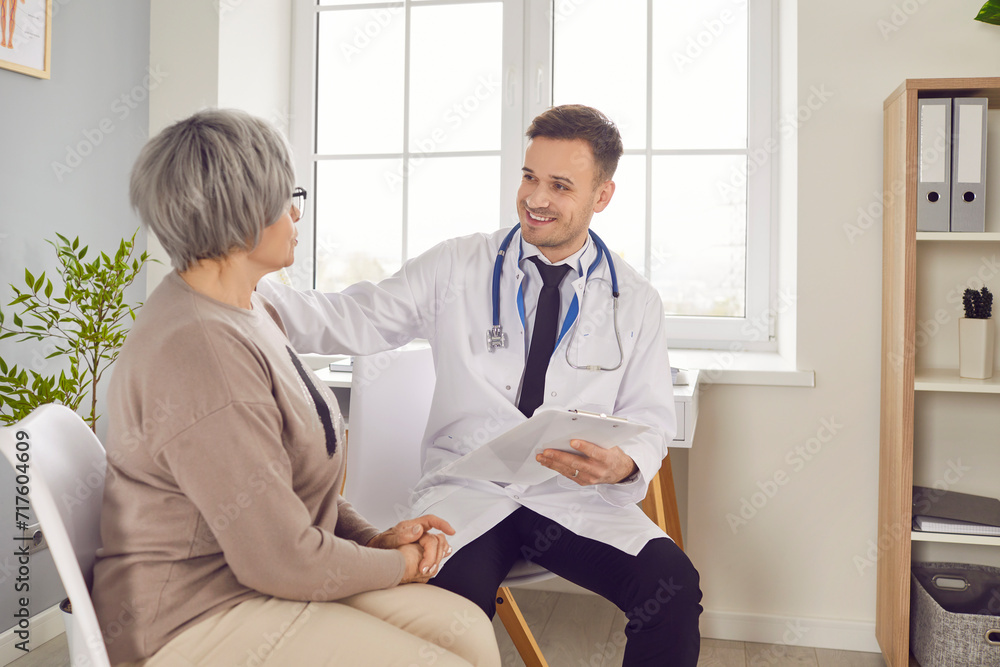 Fototapeta premium Young smiling doctor clinic visit, male nurse taking care of old woman during regular check, examination. Man monitoring resident of nursing home, patient in hospital, maintain elderly health, safety