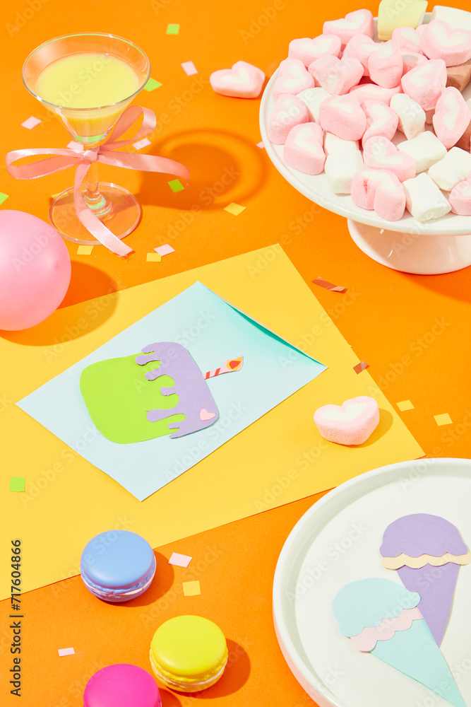 A plate of marshmallows, a cocktail, Ice cream and a birthday cake cut from colored paper, balloons and macarons on an orange background. A plate of marshmallows, a cocktail, Ice cream and a birthday cake cut from colored paper, balloons and macarons on an orange background.