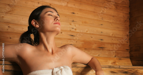 Young woman in towel relaxing in wooden sauna at spa. Asian woman in bathrobe doing body treatment in sauna.