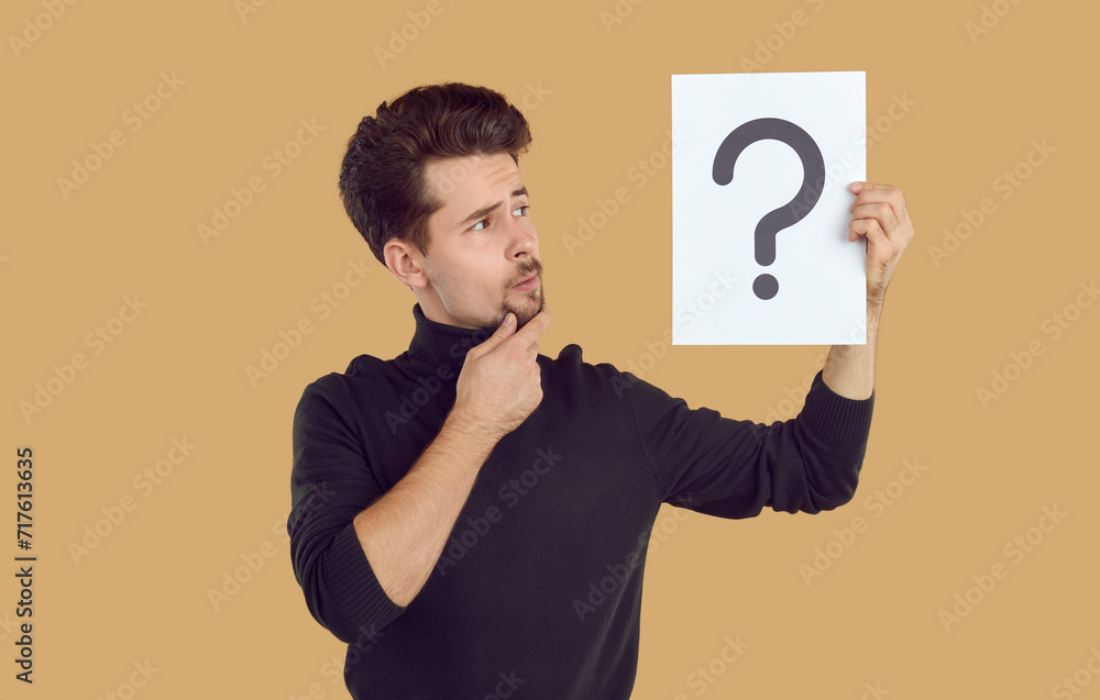 Pensive man is looking at piece of paper with question mark drawn on it, which he is holding in his hand. Young Caucasian man holding question mark symbol and thinking isolated on beige background. Pensive man is looking at piece of paper with question mark drawn on it, which he is holding in his hand. Young Caucasian man holding question mark symbol and thinking isolated on beige background.