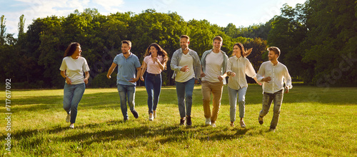 Cheerful young men and women having fun running on green lawn in park on sunny summer day. Concept of people and friendship. Friends in comfortable casual clothes having fun outdoors. Panoramic banner photo