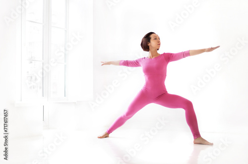 Young slender fit woman in pink long bodysuit performing yoga pose, isolated over white studio background