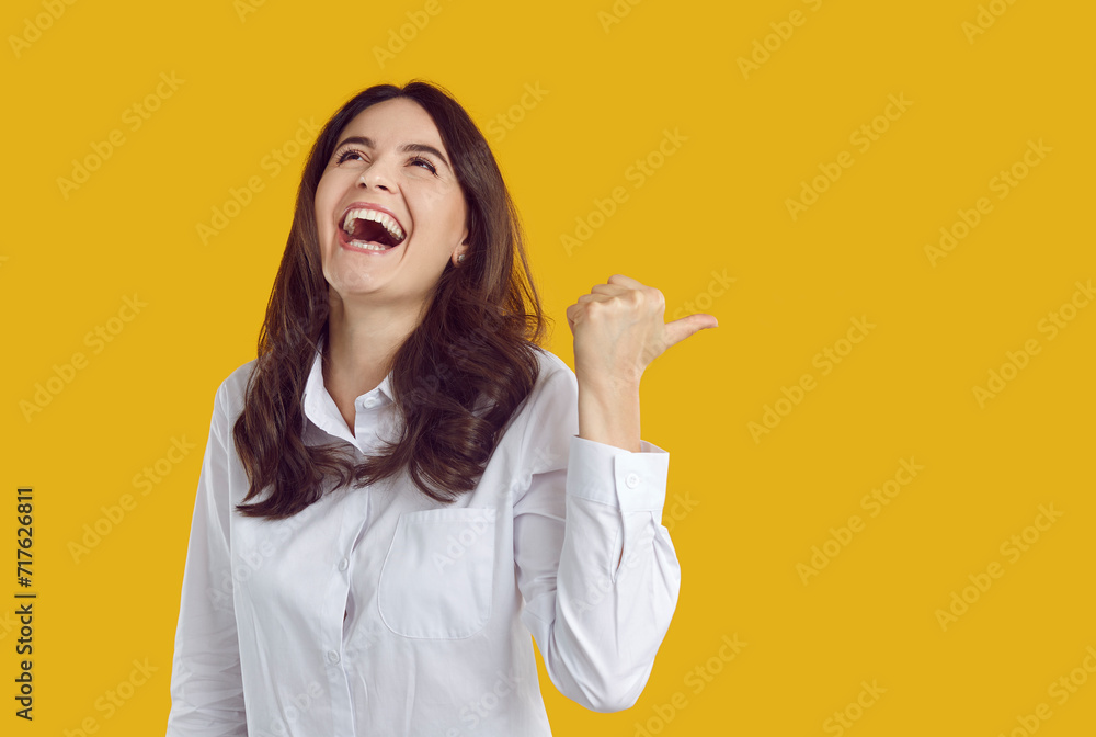 Happy laughing young woman pointing to side with thumb up. Brunette girl wearing white shirt happily smiling looking up over yellow isolated background with copy space. Positive emotions, good mood Happy laughing young woman pointing to side with thumb up. Brunette girl wearing white shirt happily smiling looking up over yellow isolated background with copy space. Positive emotions, good mood