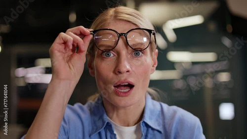 Amazed woman in casual outfit staring at something unbelievable on laptop screen at workplace. Pretty female lifting glasses with expression of pleasant surprise and satisfaction with excellent news.