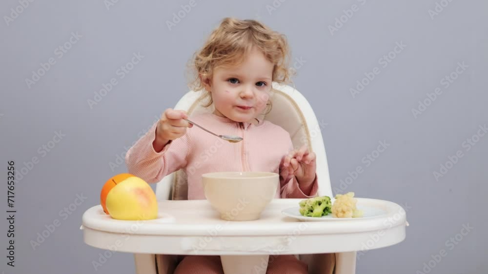 Cultivating healthy eating habits. Tasty baby dishes. Infant's healthy nutrition. Funny little baby girl with wavy hair eating in highchair isolated over gray background Cultivating healthy eating habits. Tasty baby dishes. Infant's healthy nutrition. Funny little baby girl with wavy hair eating in highchair isolated over gray background