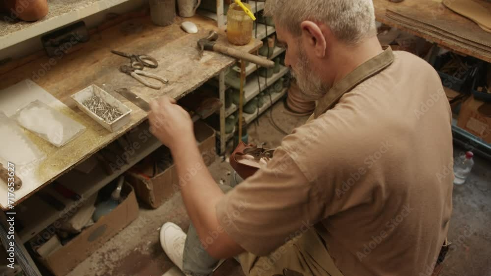 High angle view of senior artisan sitting in shoemaking workshop and attaching leather to sole with plies High angle view of senior artisan sitting in shoemaking workshop and attaching leather to sole with plies