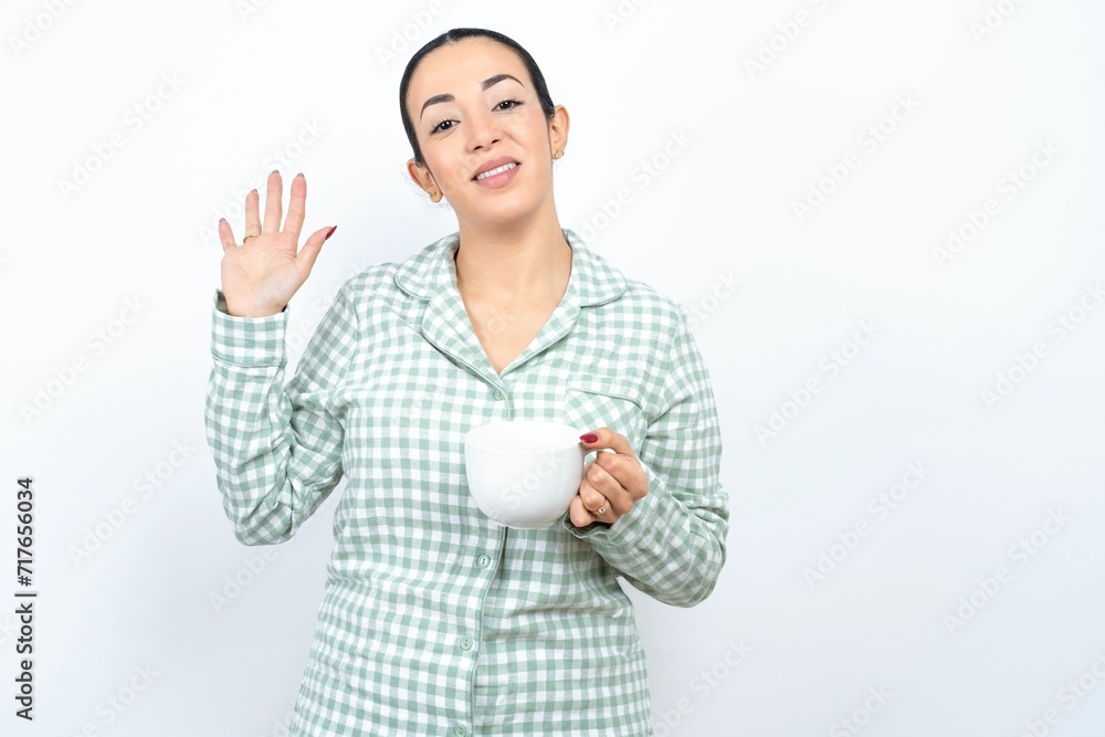 Beautiful young woman wearing green plaid pyjama and holding a cup Waiving saying hello happy and smiling, friendly welcome gesture. Beautiful young woman wearing green plaid pyjama and holding a cup Waiving saying hello happy and smiling, friendly welcome gesture.