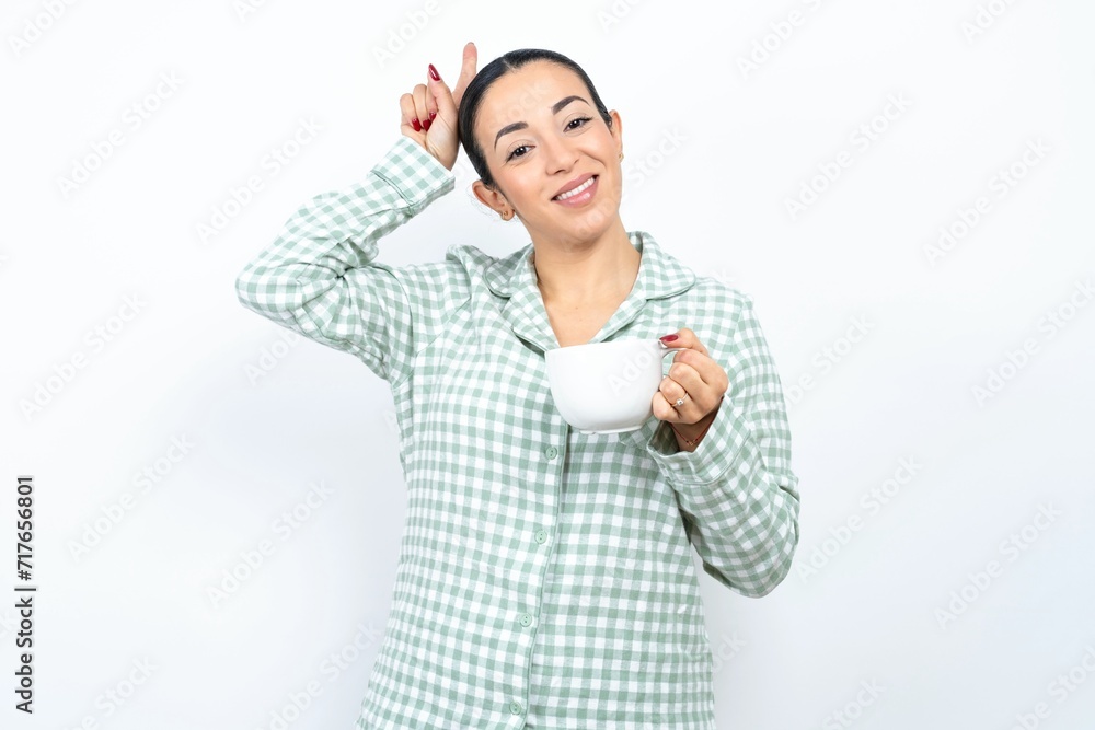 Funny Beautiful young woman wearing green plaid pyjama and holding a cup shows horns, fingers on head gesture, posing silly and cute Funny Beautiful young woman wearing green plaid pyjama and holding a cup shows horns, fingers on head gesture, posing silly and cute