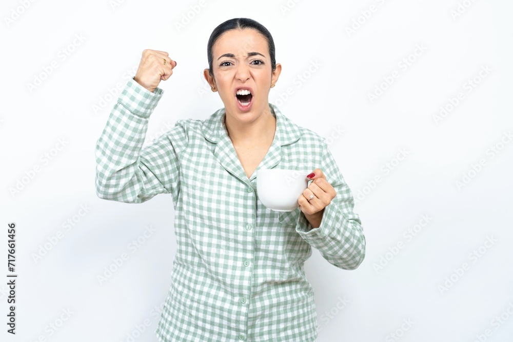 Beautiful young woman wearing green plaid pyjama angry and mad raising fist frustrated and furious while shouting with anger. Rage and aggressive concept. Beautiful young woman wearing green plaid pyjama angry and mad raising fist frustrated and furious while shouting with anger. Rage and aggressive concept.