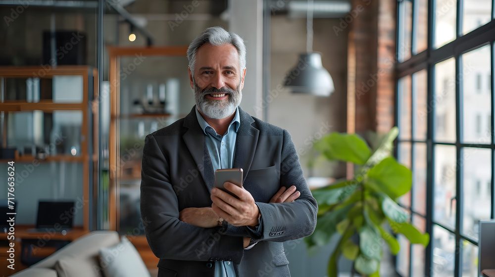 An elderly executive with a smile, uses a mobile phone while standing in his office. Enjoying a happy middle-aged career as a professional manager while using a smartphone to check financial transacti