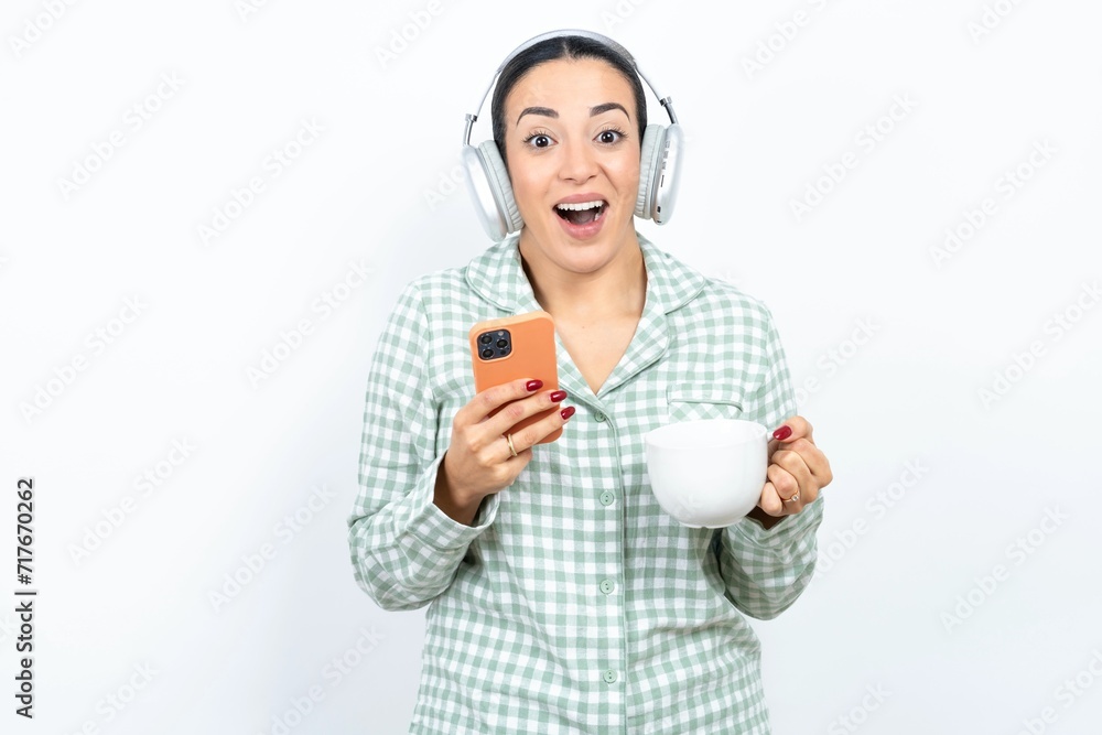 Excited Beautiful young woman wearing green plaid pyjama holding smartphone and looking amazed to the camera after receiving good news. Excited Beautiful young woman wearing green plaid pyjama holding smartphone and looking amazed to the camera after receiving good news.