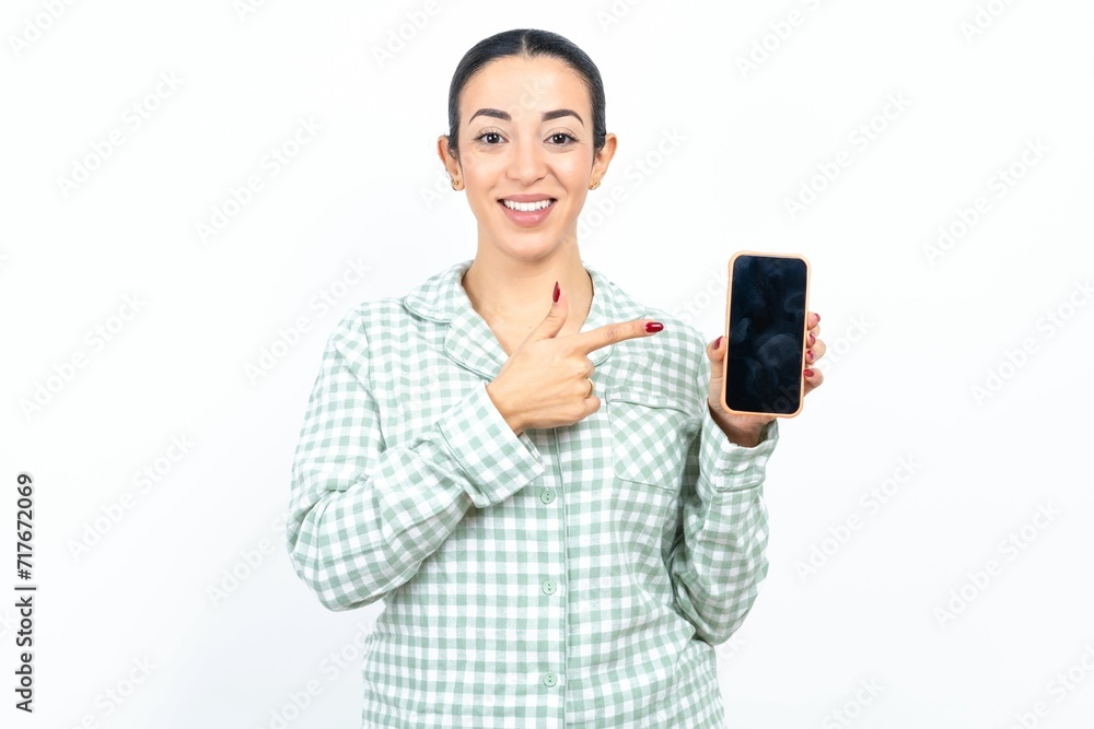 Smiling Beautiful young woman wearing green plaid pyjama Mock up copy space. Pointing index finger on mobile phone with blank empty screen Smiling Beautiful young woman wearing green plaid pyjama Mock up copy space. Pointing index finger on mobile phone with blank empty screen
