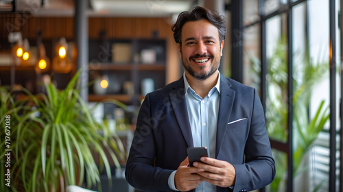 Smiling and content, a young Latino manager or CEO stands in an office, holding a smartphone and uses it to make a bank transfer while averting his eyes from his work photo
