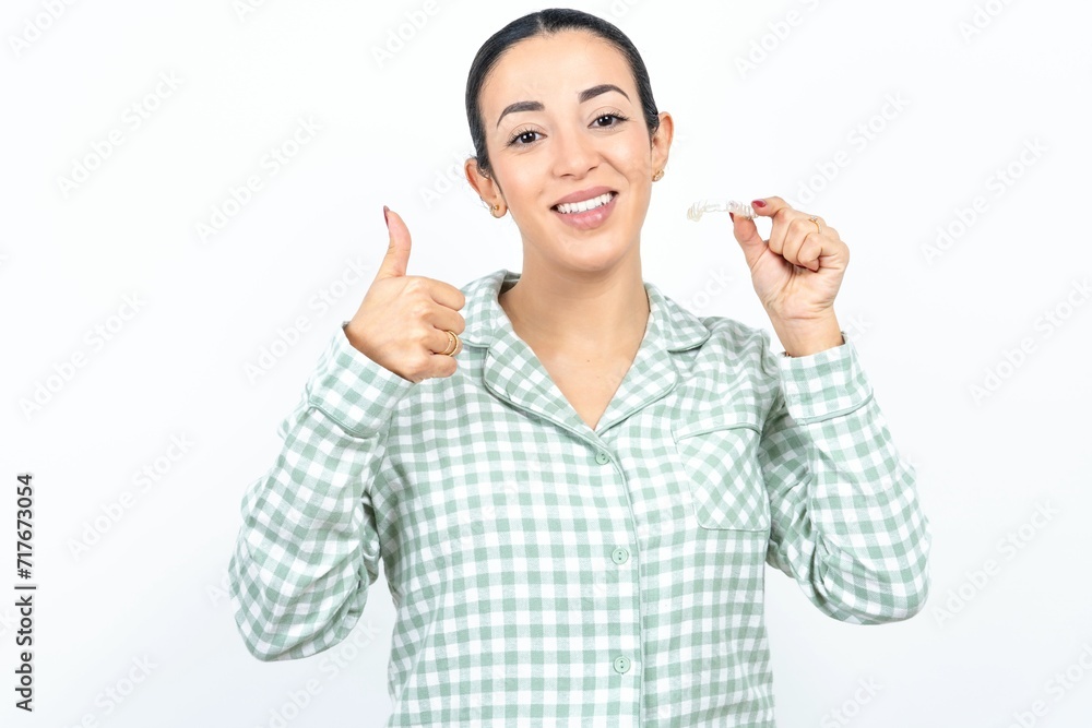 Beautiful young woman wearing green plaid pyjama holding an invisible braces aligner and rising thumb up, recommending this new treatment. Dental healthcare concept. Beautiful young woman wearing green plaid pyjama holding an invisible braces aligner and rising thumb up, recommending this new treatment. Dental healthcare concept.
