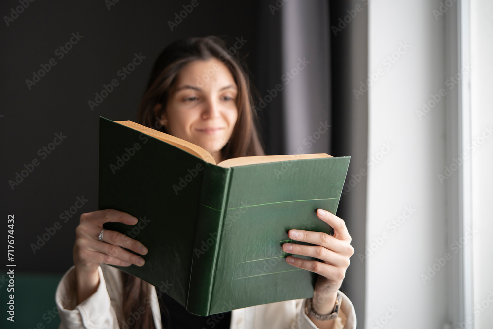 A woman stands by a large window, bathed in natural light, deeply focused on reading an open book. A woman stands by a large window, bathed in natural light, deeply focused on reading an open book.
